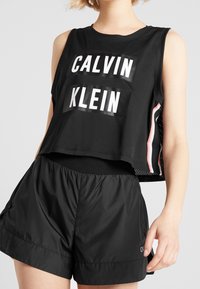 Mustat lyhyet topit, joissa on valkoinen "CALVIN KLEIN" -logo, yhdistettyinä mustiin kevyisiin shortseihin. Topissa on verkkokankaisia sivupaneeleja ja raidallisia yksityiskohtia.