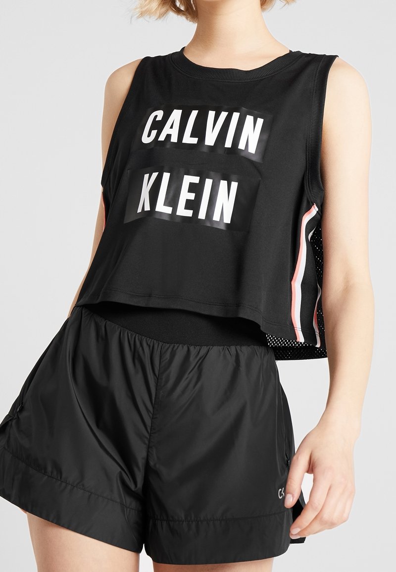 Mustat lyhyet topit, joissa on valkoinen "CALVIN KLEIN" -logo, yhdistettyinä mustiin kevyisiin shortseihin. Topissa on verkkokankaisia sivupaneeleja ja raidallisia yksityiskohtia.