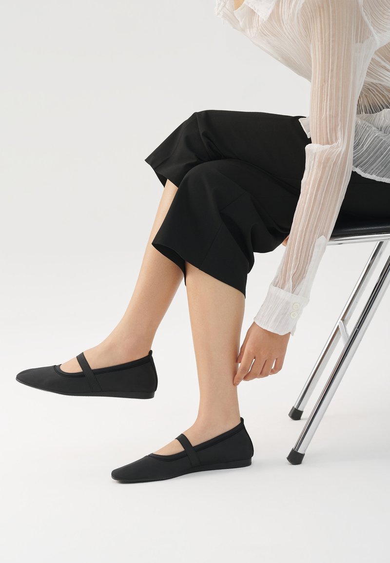 Ballerine nere con punta arrotondata, dotate di una fascia sulla parte superiore del piede, realizzate in materiale liscio con una leggera texture.