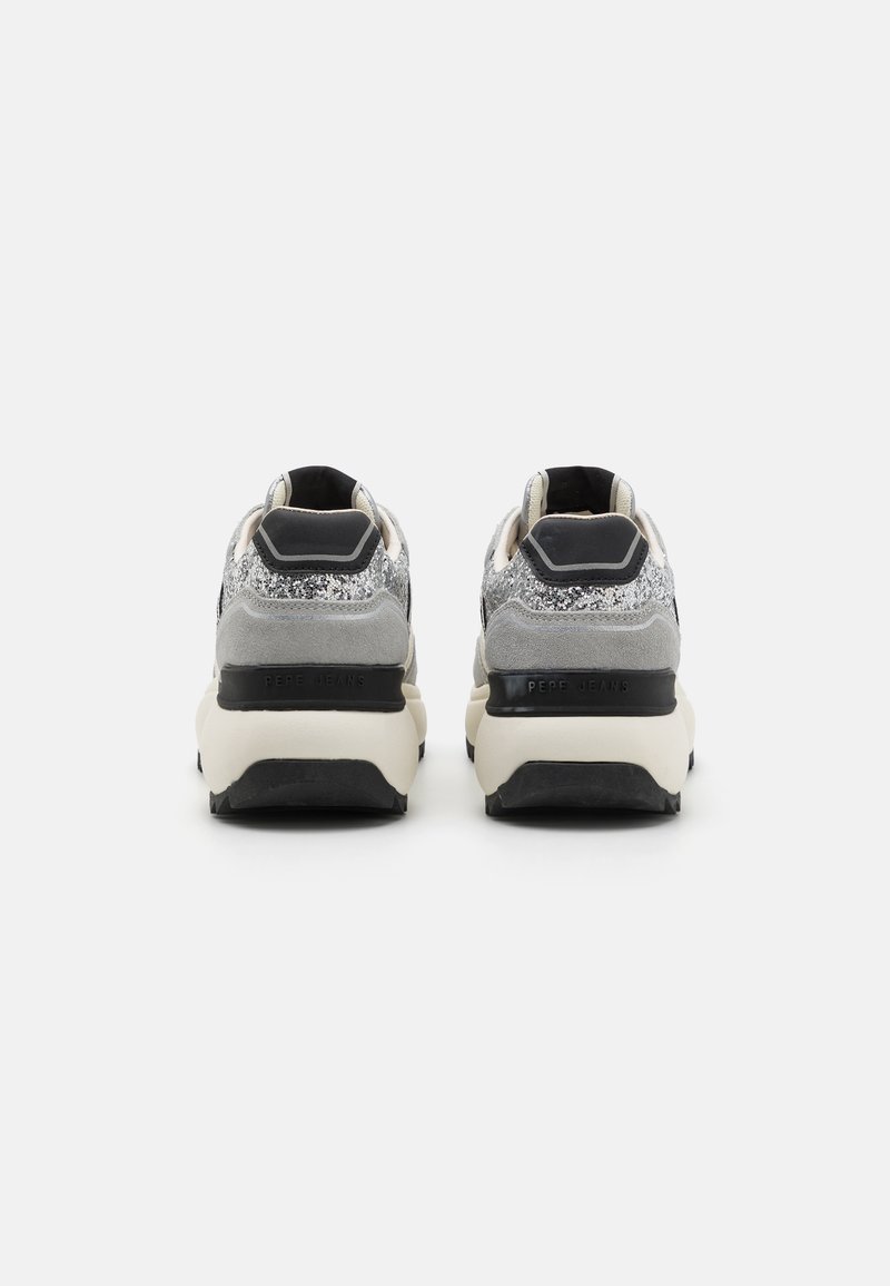 Vista trasera de unas zapatillas que presentan una parte superior de glitters en gris y plata, acentos en negro, y una gruesa suela blanca con el logo de la marca y patrones texturizados.