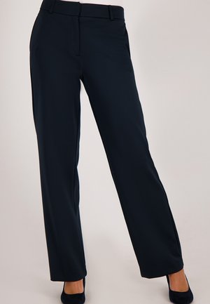 Vrouw in een rechte zwarte broek en zwarte hakken tegen een effen lichte achtergrond.