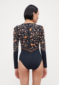 Maillot de bain à manches longues avec un motif floral orange et blanc sur un fond marine, avec une fermeture éclair au dos et un design épuré.
