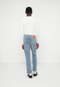 Ανοιχτού μπλε τζιν παντελόνι με ίσια γραμμή, εμφανίζονται από πίσω. Διαθέτει πορτοκαλί ετικέτα στη μέση και σκούρα παπούτσια.