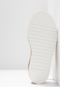 Semelle d'une chaussure à semelle blanche avec une texture striée, reposant verticalement contre un mur et une surface blanche.