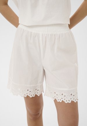 Biele bavlnené šortky s elastickým pásom a lemom z očkovanej čipky so zvlneným okrajom, ktoré nosí osoba s rukami za chrbtom.