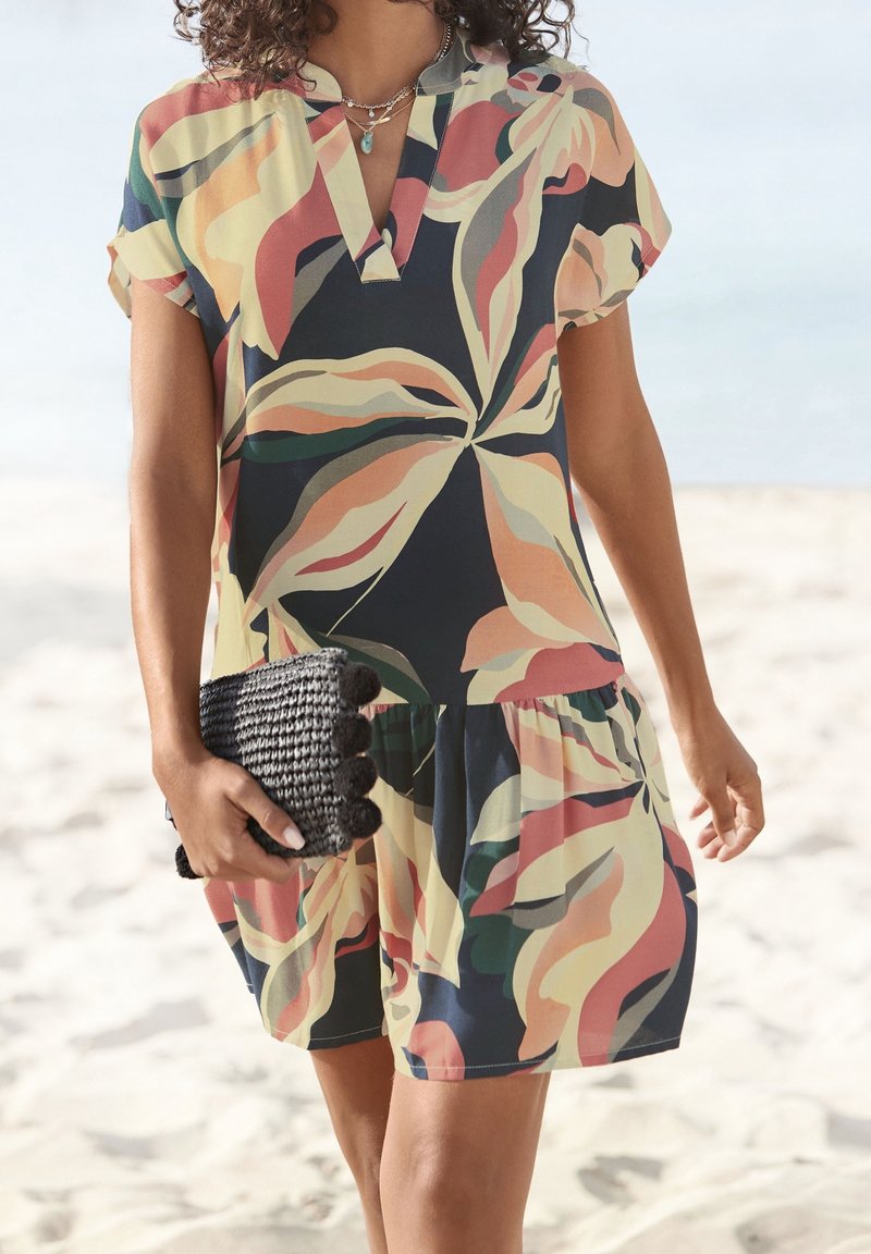 Femme portant une robe courte à manches courtes imprimée d'un motif abstrait de feuilles colorées, tenant une pochette noire tissée sur une plage de sable.