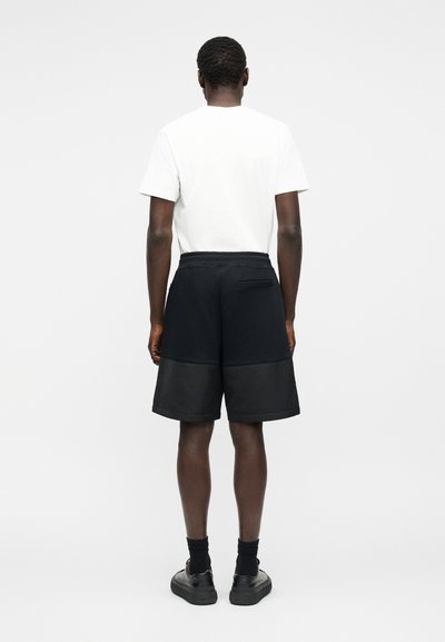 Άνδρας που φοράει λευκό πουκάμισο με κοντά μανίκια, μαύρα σορτς με τσέπη στο πίσω μέρος, μαύρες κάλτσες και μαύρα παπούτσια, στέκεται μπροστά σε απλό φόντο.