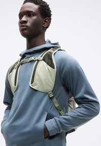 Ανοιχτό γκρι και ελιάς πράσινο σακίδιο με ρυθμιζόμενους ιμάντες, με υφή στην επιφάνεια και μεγάλες πλαϊνές τσέπες, φορεμένο πάνω από μπλε φούτερ με μακριά μανίκια.