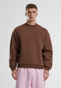 Sudadera marrón de gran tamaño con cuello redondo alto, puños y dobladillo acanalados; textura suave y corte relajado; combinada con pantalones rosa claro.