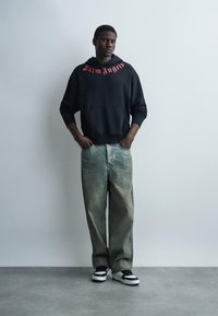 Νεαρός άνδρας στέκεται με τα χέρια στις τσέπες, φοράει μαύρη μπλούζα με κουκούλα "Palm Angels", φαρδιά ξεθωριασμένα τζιν και ασπρόμαυρα αθλητικά παπούτσια μπροστά σε απλό τοίχο.