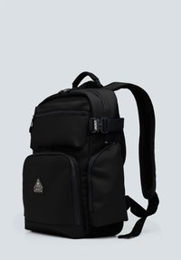 Mochila de tela negra con múltiples bolsillos con cremallera, correas de hombro acolchadas y un logo triangular en la parte frontal. Textura suave.