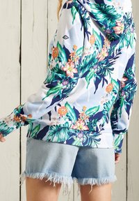 Persona que lleva una sudadera con patrón floral de hojas y flores en verde y naranja, combinada con pantalones cortos de mezclilla claros y deshilachados.