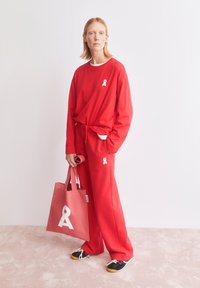 Rotes langärmeliges Oberteil mit weißem Logo, kombiniert mit passenden Hosen. Das Model hält eine pinke Tragetasche mit weißem Motiv. Sneaker vervollständigen das Outfit.