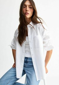 Chemise blanche à boutons avec une poche poitrine, manches retroussées et coupe décontractée, portée sur un débardeur blanc et associée à un jean bleu clair.