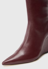 Close-up of a burgundy leather wedge boot with a pointed toe and smooth stitching on a plain background.