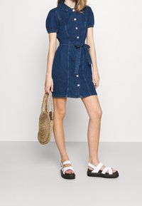 Kvinna i en kortärmad denimknäppklänning med bälte, som håller i en rund vävd handväska och bär vita plattformssandaler.