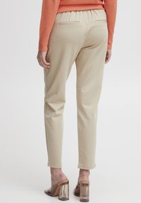 Pantalon en toile beige avec une taille élastique, doté de deux poches arrière. Porté avec des mules à talons transparents, sur un fond sobre et clair.