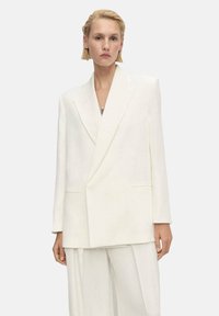 Blazer bianco oversize con lapel affilati, spalle strutturate e tasche frontali, realizzato in un tessuto liscio e texturizzato.
