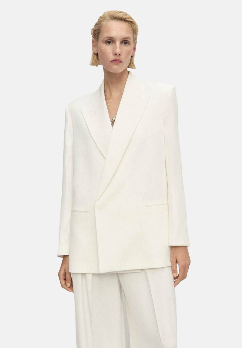 Blazer bianco oversize con lapel affilati, spalle strutturate e tasche frontali, realizzato in un tessuto liscio e texturizzato.