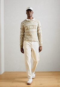 Maglione lavorato a maglia con un motivo geometrico in beige e crema, abbinato a pantaloni beige chiaro e sneakers bianche. Il modello indossa un cappellino bianco.