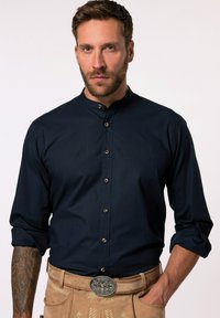 Camicia a maniche lunghe blu navy con colletto alto e bottoni dorati, abbinata a lederhosen color crema con una fibbia metallica ornata.