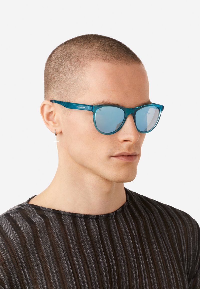 Un par de gafas de sol azules con lentes espejadas, con una montura transparente y un diseño elegante, usadas por un modelo con cabello corto.