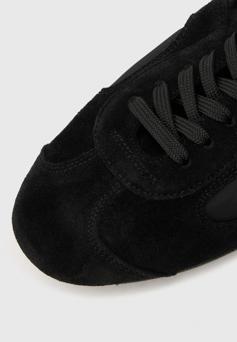 Bout de basket en daim noir avec une forme arrondie, des détails cousus et des lacets noirs tissés passés à travers des œillets.