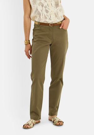 Femme portant un pantalon vert olive, un haut en crochet beige, une ceinture marron, des bracelets dorés et des sandales dorées à bouts ouverts, debout avec une main dans la poche.