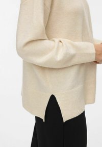 Bež pulover iz pletene tkanine z rebrastimi manšetami in robom, z stransko odprtino, teksturirano tkanino in sproščenim fitom. V kombinaciji s črnimi hlačami.
