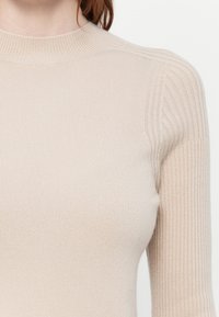 Nærbilde av en person som har på seg en beige ribbestrikket genser med høy hals og lange ermer.