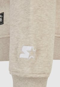 Manche de sweat-shirt beige avec un "S" blanc brodé et un logo étoile au-dessus du bord-côte côtelé, devant présente un patch rectangulaire noir avec une étoile blanche.