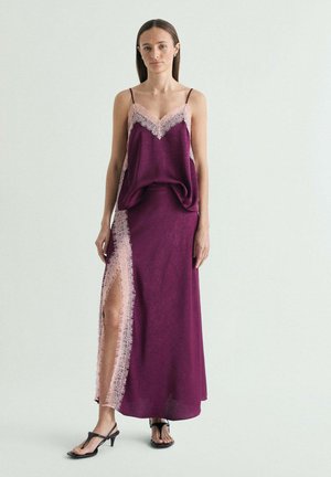 Mujer de pie con un vestido lencero morado sin mangas con ribete de encaje rosa y una abertura hasta el muslo, combinado con sandalias negras de tacón.