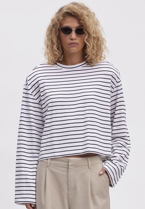 Cămașă albă cu mâneci lungi, croită pe jumătate, cu dungi orizontale negre, dintr-un material de bumbac moale și guler rotund, completată cu pantaloni bej.