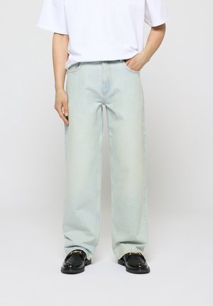 Personne portant un jean large bleu clair avec une main dans la poche et des mocassins noirs avec des détails en chaîne dorée, sur un fond uni.