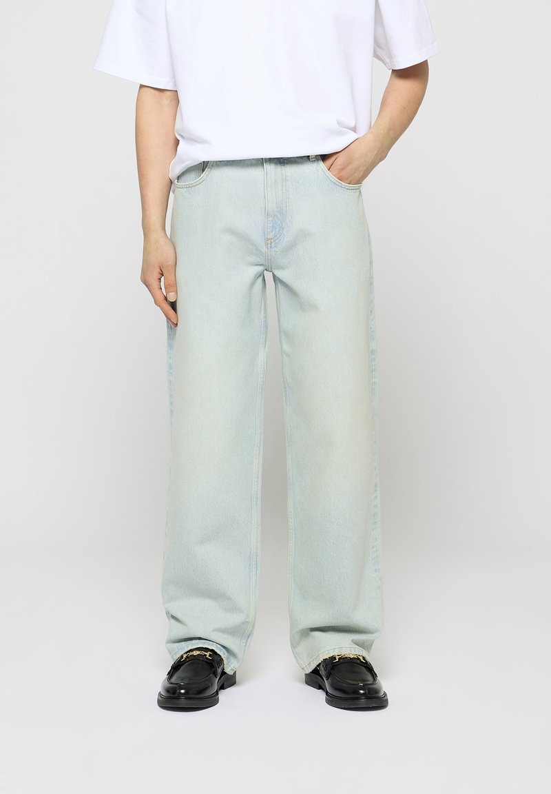 Personne portant un jean large bleu clair avec une main dans la poche et des mocassins noirs avec des détails en chaîne dorée, sur un fond uni.
