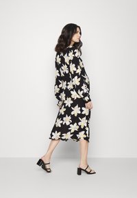 Vestido preto floral com flores amarelas e brancas, mangas longas e comprimento médio. Usado com sandálias de bloco de salto pretas com tiras.