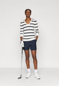 Marineblå og hvite stripegenser med v-hals, ribbet tekstur, kombinert med marineblå shorts. Modellen holder en golfklubbe og har på seg hvite sportssko.