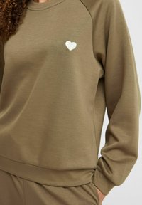 Sudadera verde oliva con cuello redondo acanalado, mangas raglán y pequeño bordado de corazón blanco en el lado izquierdo del pecho. Textura suave.