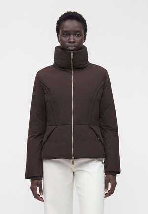 Frau trägt eine braune Jacke mit hohem Kragen und Reißverschluss sowie weiße Hose, steht vor hellem schlichten Hintergrund und schaut geradeaus.