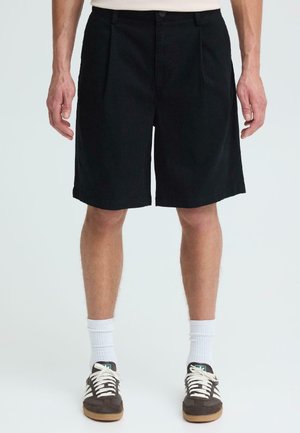 Mężczyzna ubrany w czarne krótkie spodenki za kolano, białe skarpetki i brązowe trampki z białymi sznurowadłami, stojący na jasnym, jednolitym tle.