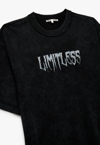Czarna bawełniana koszulka z krótkim rękawem, z teksturowanym napisem "LIMITLESS" w zużytej bieli na środku oraz ściągaczem przy dekolcie.