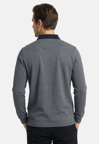 Mann mit dem Rücken zur Kamera, trägt ein hellgraues Poloshirt mit langen Ärmeln, dunklem Kragen und schwarze Hose, Hände in den Taschen.