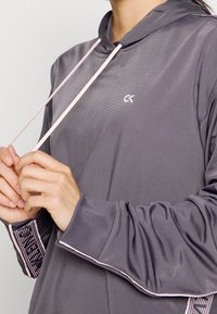 Gray athletic pullover with a hood, featuring pink drawstrings, ribbed cuffs, and patterned accents at the sleeve openings. CK logo on chest.