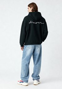 Sudadera negra con capucha que presenta el texto blanco "Amour" en la parte posterior, combinada con unos vaqueros anchos de color azul claro y zapatillas blancas con detalles en rojo y azul.