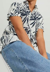 Camicia a maniche corte con bottoni, bianca con stampa botanica blu, abbinata a jeans di denim azzurro chiaro. Presenta una vestibilità comoda e colletto.