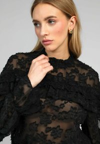 Blouse noire en voile transparent avec broderie florale texturée, accents volants, col montant et manches longues. La transparence subtile ajoute de la profondeur.
