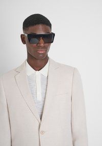Blazer en lin beige sur un polo gris avec un col crème. Des lunettes de soleil noires surdimensionnées complètent le look. Conception minimaliste, texture lisse.