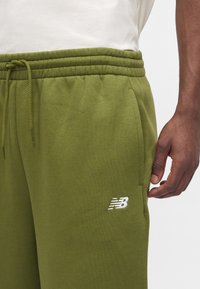 Άνδρας που φοράει χακί πράσινη φόρμα New Balance με ελαστική μέση και κορδόνι, το χέρι του ακουμπά κοντά στην πλαϊνή τσέπη.