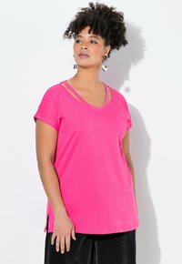 Růžové tričko s krátkým rukávem a výstřihem do V, se bočními rozparky a vrstvenými ramínky, vyrobené z měkké tkaniny. Kombinováno s černými plisovanými kalhotami.