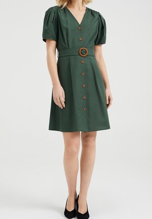 Robe verte à encolure en V avec manches bouffantes, boutons marron sur le devant, ceinture assortie avec boucle ronde, portée avec des talons pointus noirs.